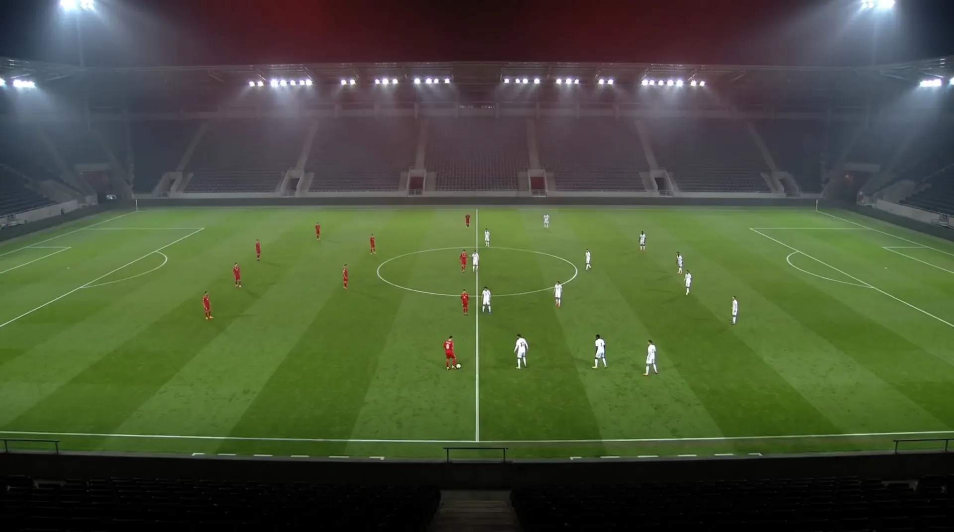 High tactical-camera view of a full 11-a-side match, red against white, under floodlights